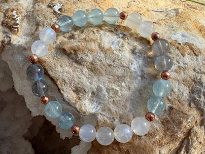 A beaded stretch bracelet made of light blue, smoky gray, and milky white round beads separated by small metallic copper spacer beads, displayed on a textured natural rock surface.