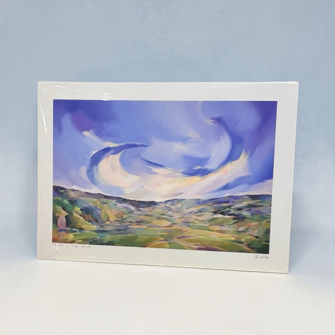 A framed landscape art print showing a stylized blue sky and green valley, photographed against a light blue background.