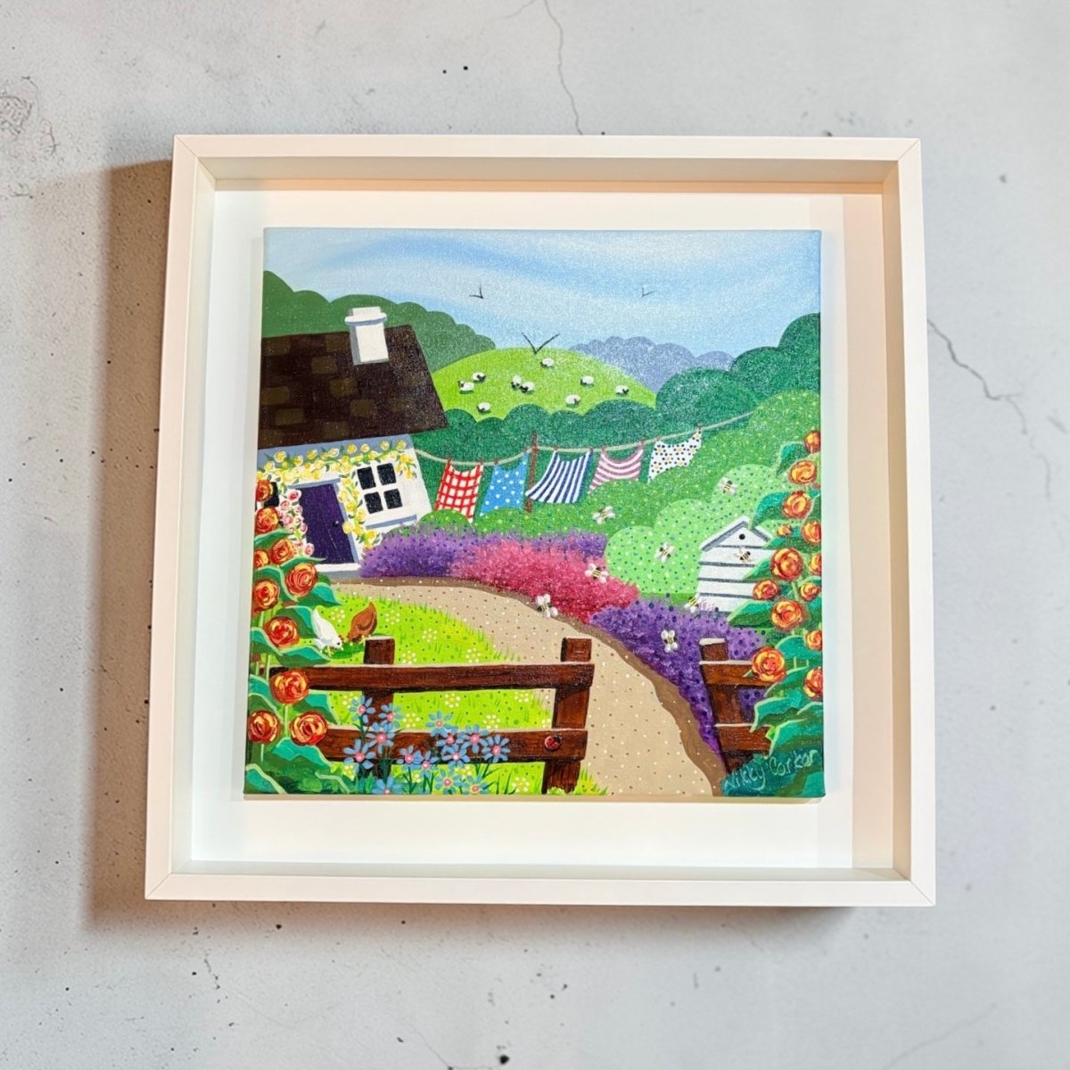 Framed artwork of a colorful village scene with a house, garden, and laundry hanging on a line.
