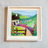 Framed artwork of a colorful village scene with a house, garden, and laundry hanging on a line.