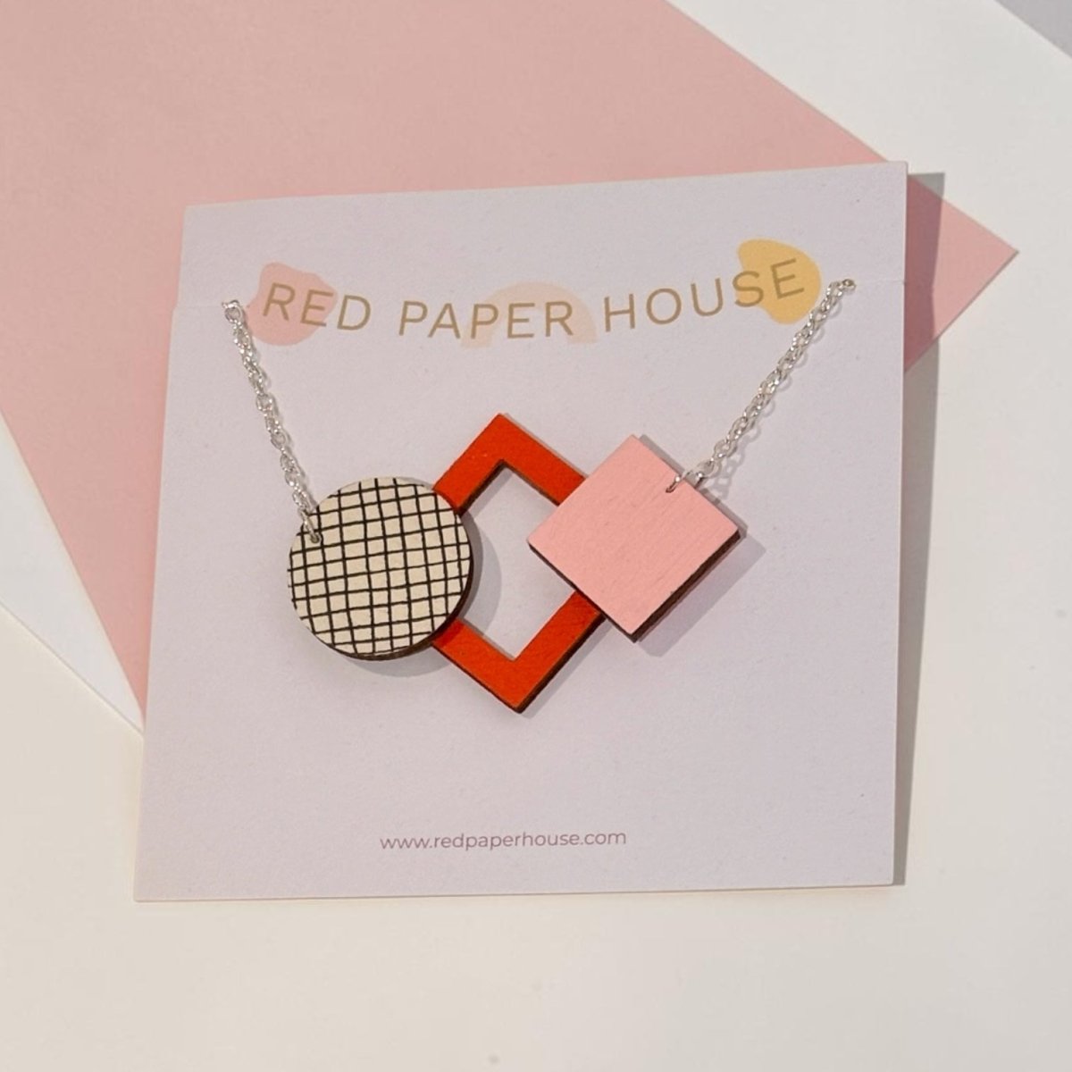 A geometric acrylic pendant necklace by Red Paper House, featuring a pink square, a red-orange diamond frame, and a black-and-white checkered circle, displayed on its branded card.