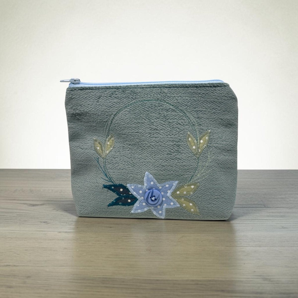 A small zippered pouch with a textured gray-green fabric body, featuring a decorative embroidered design of a blue and white star, gold leaves, and a blue ribbon on a light wood surface against a neutral background.
