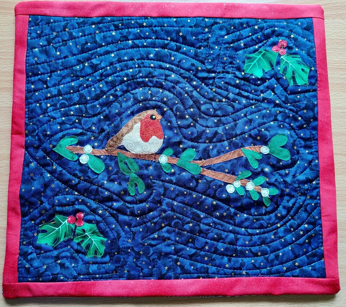 A square quilted fabric piece featuring a robin bird on a branch with mistletoe, set against a deep blue wavy background with small gold star-like details and a bright pink border.