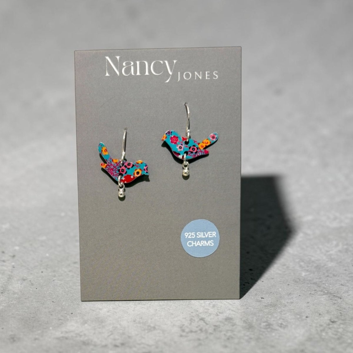 Nancy Jones bird-shaped earrings with a colorful floral pattern and silver hooks, displayed on a grey brand card against a textured grey background.