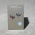 Nancy Jones bird-shaped earrings with a colorful floral pattern and silver hooks, displayed on a grey brand card against a textured grey background.