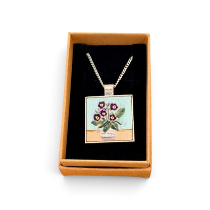 A square pendant necklace displayed inside an open tan gift box with a black insert. The pendant shows a floral bouquet in a vase with purple flowers and green leaves on a light blue and beige background, suspended from a silver-toned chain.