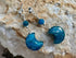A pair of dangle earrings with polished blue crescent moon charms, blue faceted beads, and silver-toned metal hooks, displayed on a textured beige rock surface.