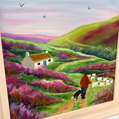 A framed landscape painting depicting rolling green hills, pink-purple heather fields, a small white cottage with a brown roof, sheep, a shepherd in a red jacket, a black dog, and birds in a pastel sky.
