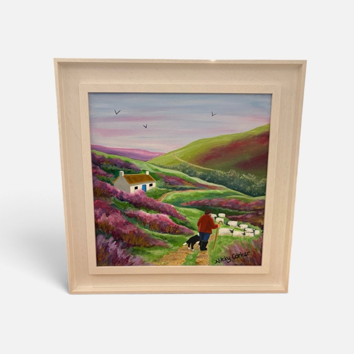 A framed landscape painting on a light gray background. The artwork depicts rolling green hills, purple heather, a small white cottage, and a figure walking with a dog.