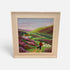 A framed landscape painting on a light gray background. The artwork depicts rolling green hills, purple heather, a small white cottage, and a figure walking with a dog.