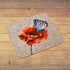 A square cork placemat with rounded corners featuring a printed design of a blue butterfly resting on a red-orange poppy flower, displayed on a rustic wooden plank surface.