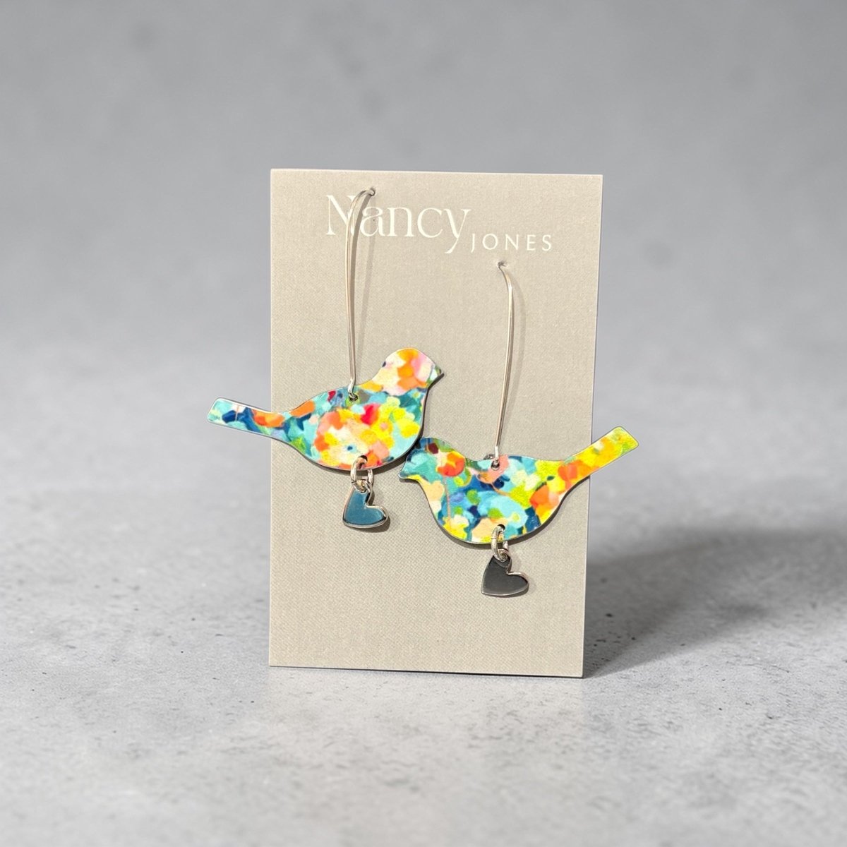 Colorful bird-shaped earrings on a Nancy Jones card against a gray background