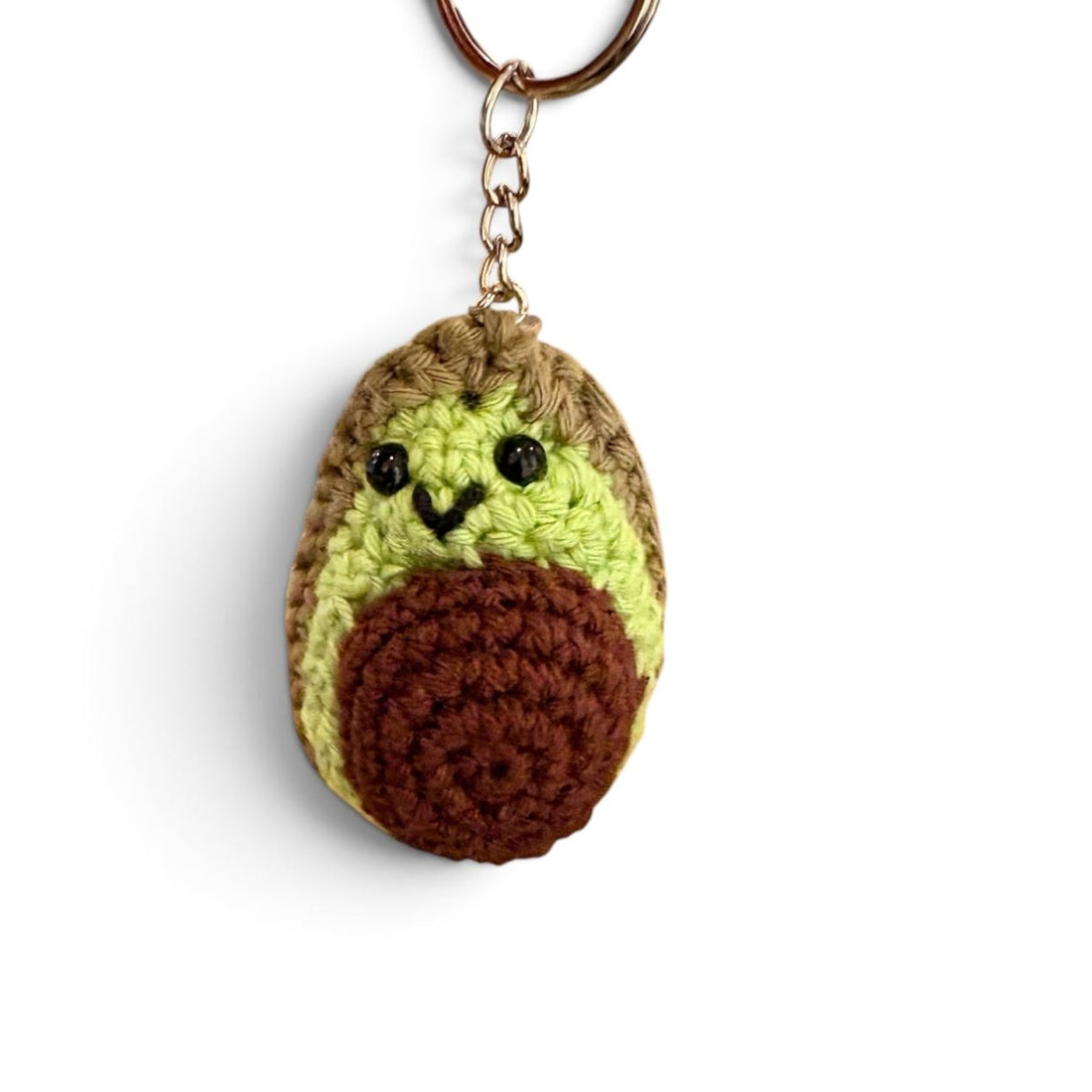 A crocheted avocado keychain featuring a light green textured body, a brown textured skin, embroidered black eyes and mouth, and a gold metal keyring at the top, shown on a plain white background.