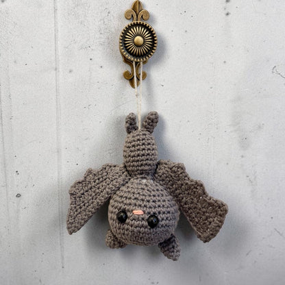 A crocheted bat hanging ornament in a medium gray color, featuring black embroidered eyes and a small pink nose, suspended from an antique bronze wall hook against a textured gray wall.