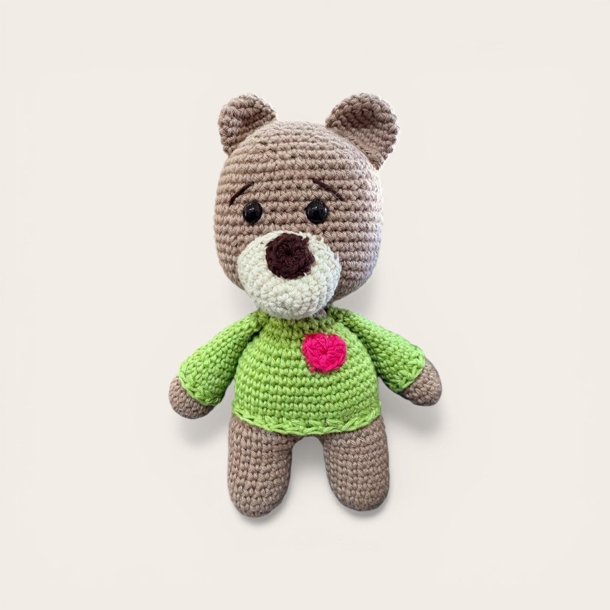 Crocheted teddy bear with a green sweater and pink heart on a light beige background