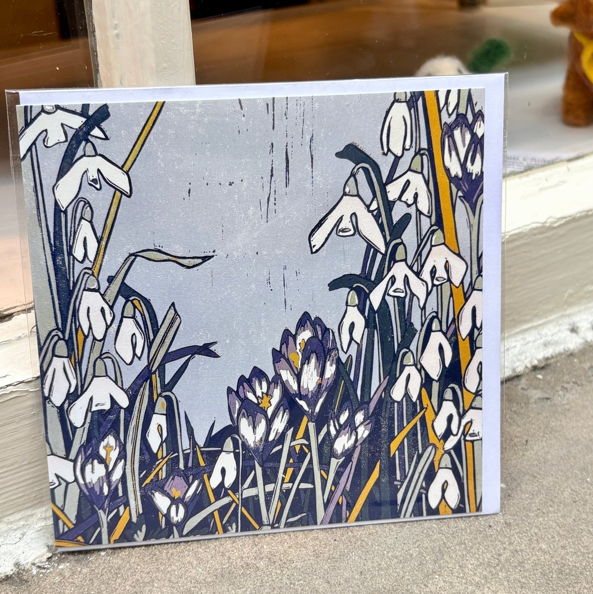 A square art card featuring a stylized floral illustration of snowdrops and crocuses in white, purple, navy, gray, and mustard yellow, leaning against a window ledge.