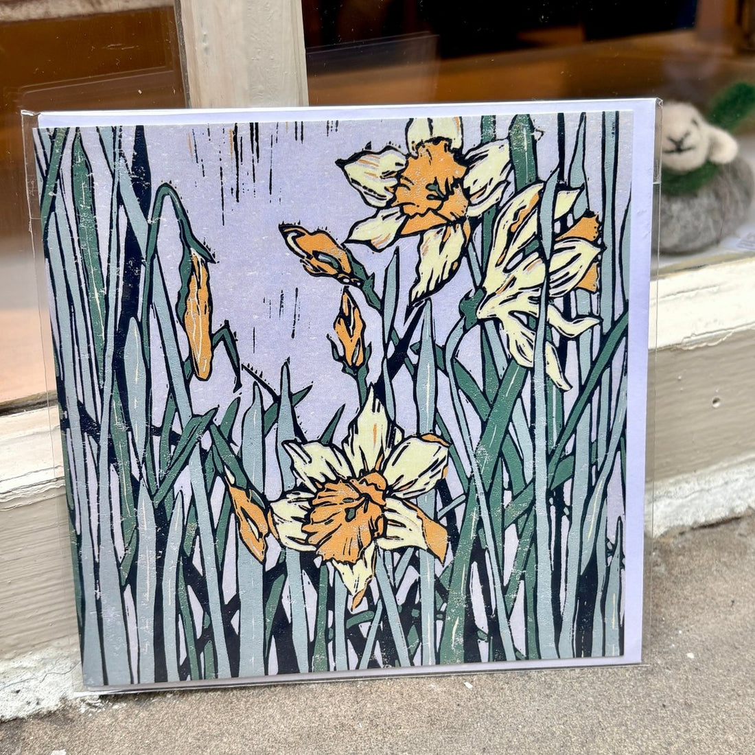 A square art print or card featuring a stylized floral illustration of daffodils and tall leaves, photographed leaning against a window or door frame outdoors.