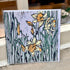 A square art print or card featuring a stylized floral illustration of daffodils and tall leaves, photographed leaning against a window or door frame outdoors.