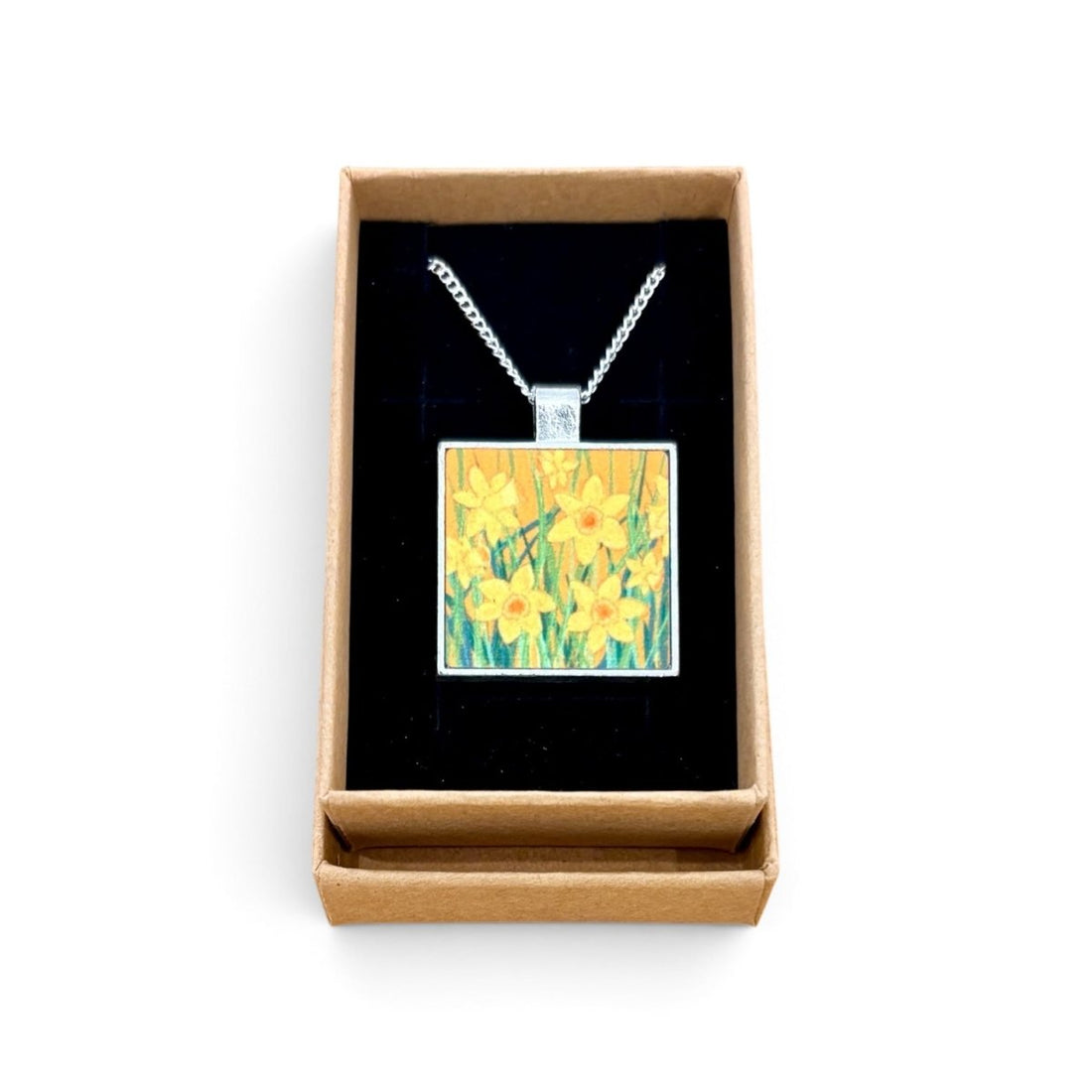 A square pendant necklace with a daffodil painting design, displayed inside an open brown gift box with a black velvet lining on a white background.