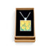 A square pendant necklace with a daffodil painting design, displayed inside an open brown gift box with a black velvet lining on a white background.