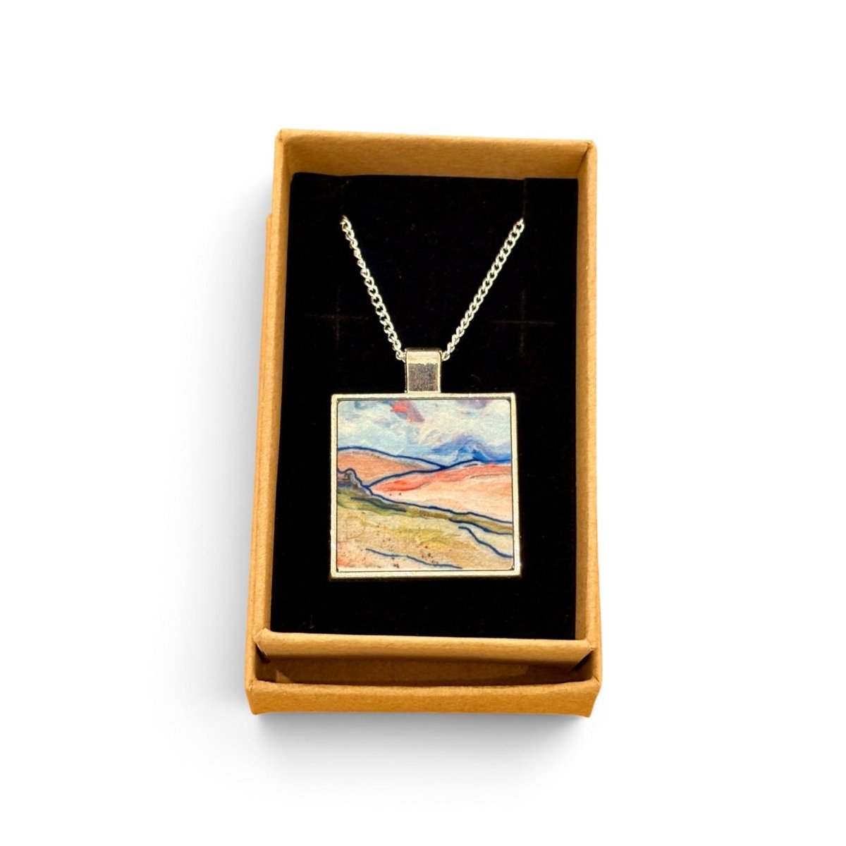 A square pendant necklace displayed in an open tan gift box with a black insert. The pendant features a colorful landscape painting with blue, pink, yellow, and green tones, suspended from a silver-toned chain.