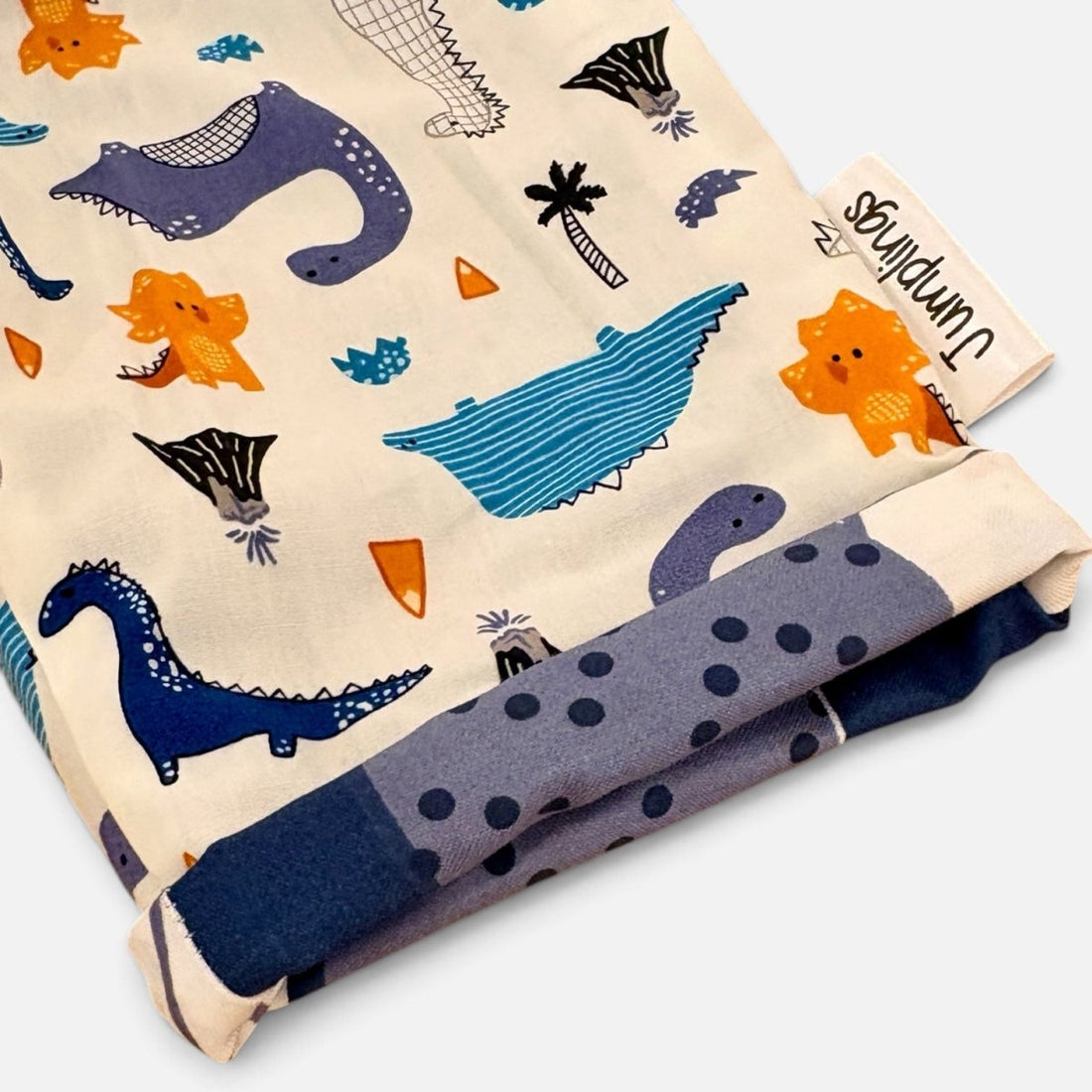 A folded fabric item, likely a blanket or scarf, featuring a whimsical dinosaur and prehistoric animal print on a cream background. The print includes blue, purple, and orange dinosaurs, alongside a matching blue polka-dot reverse side.