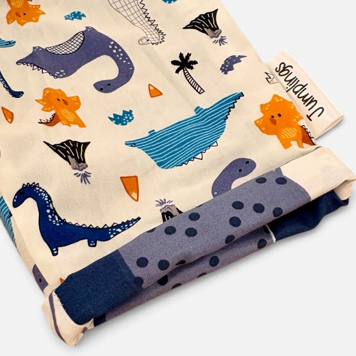A folded fabric item, likely a blanket or scarf, featuring a whimsical dinosaur and prehistoric animal print on a cream background. The print includes blue, purple, and orange dinosaurs, alongside a matching blue polka-dot reverse side.