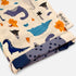 A folded fabric item, likely a blanket or scarf, featuring a whimsical dinosaur and prehistoric animal print on a cream background. The print includes blue, purple, and orange dinosaurs, alongside a matching blue polka-dot reverse side.