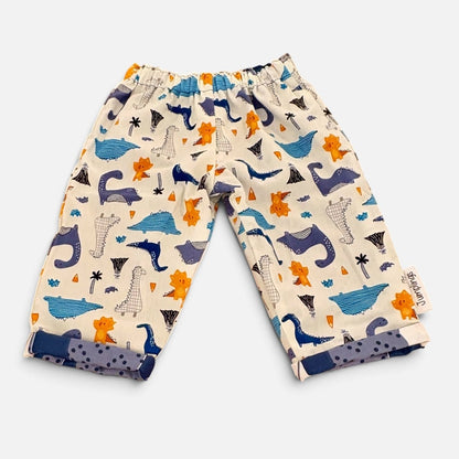 A pair of baby pants with an elastic waistband, featuring a playful dinosaur print in navy blue, orange, and gray on a cream-colored background. The pants are shown flat-laid on a light gray background.
