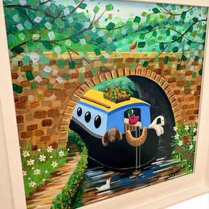 A colorful framed painting depicting a blue and yellow canal house on water under a brick bridge, surrounded by green foliage and small white flowers. The artwork is shown inside a light wood frame.