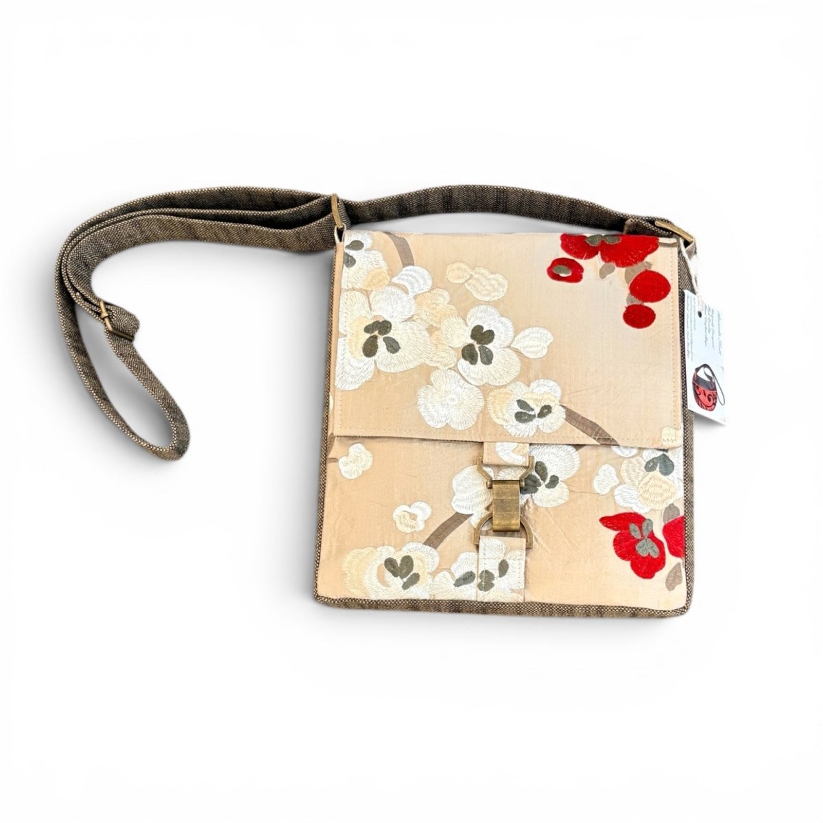 A small crossbody bag with a beige floral print, featuring white blossoms, red flowers, and a brown strap, shown on a white background with a product tag attached.