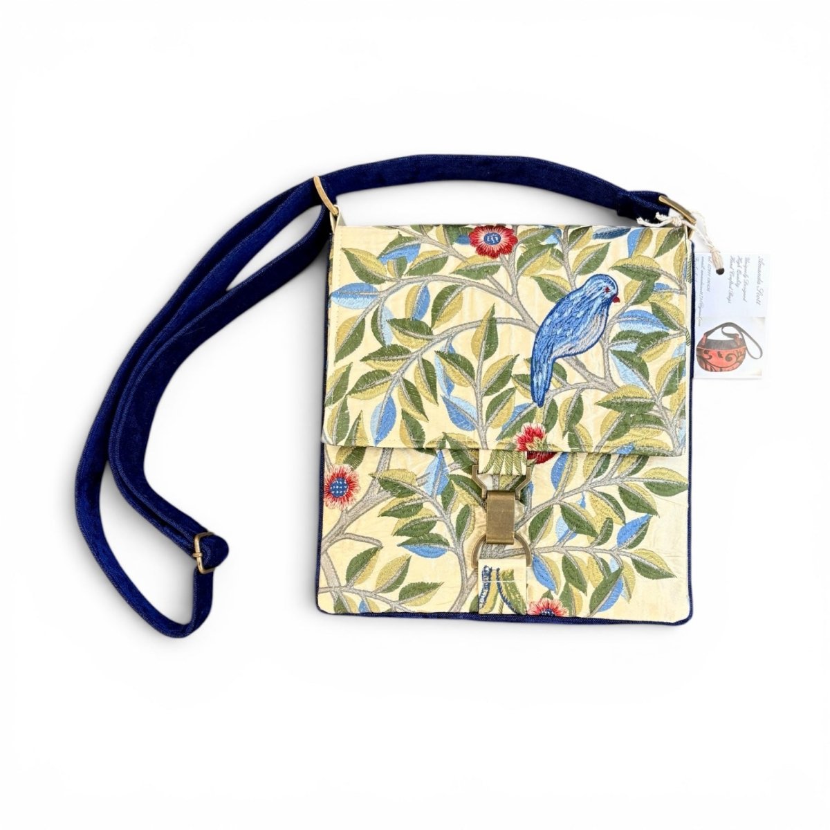 A small crossbody handbag with a navy blue strap and a front flap featuring a William Morris-style floral pattern in green, blue, red, and cream. A small hanging tag is attached on the right side.