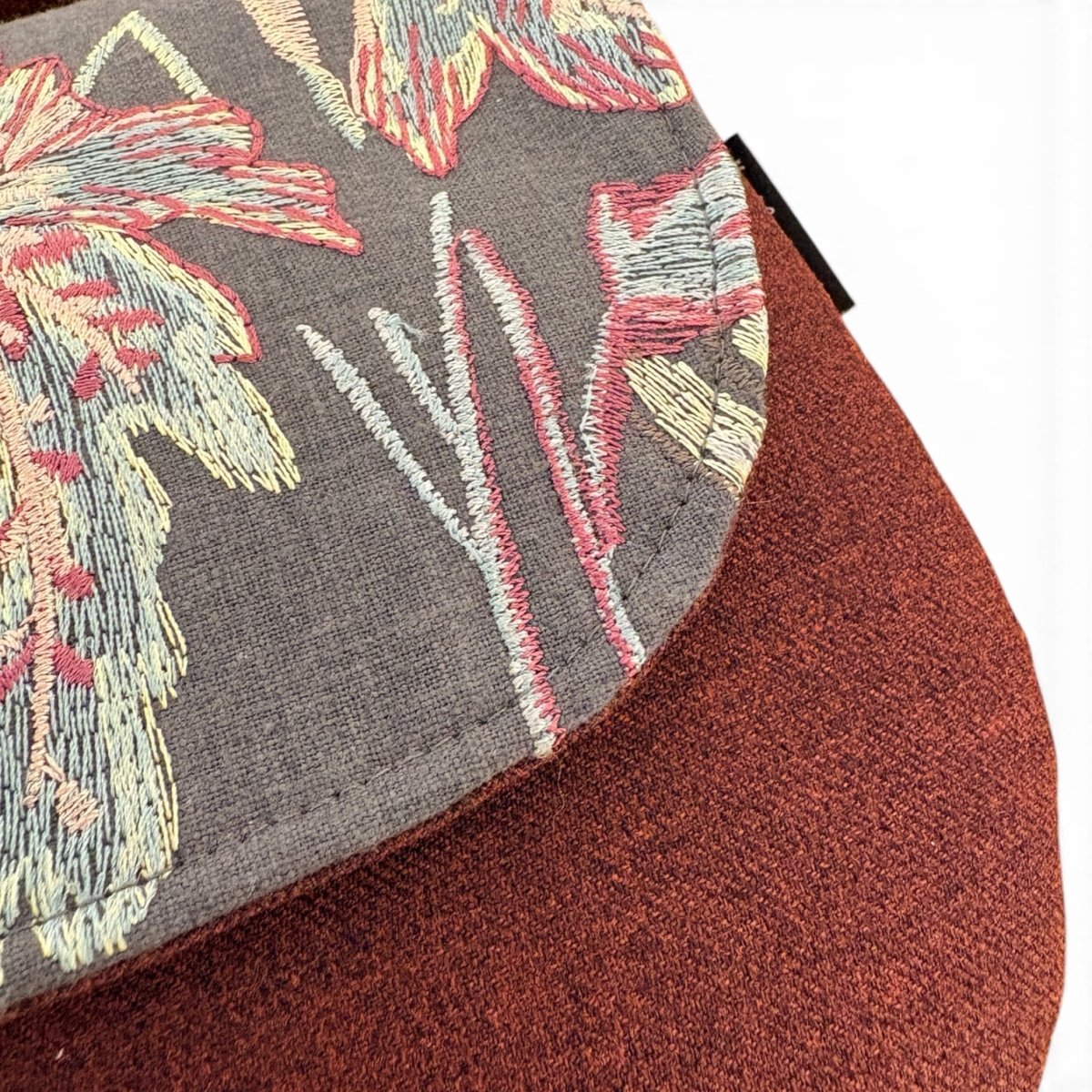 Close-up of a cap showing a dark gray embroidered floral patterned fabric and a rust-brown textured fabric side, set against a plain white background.