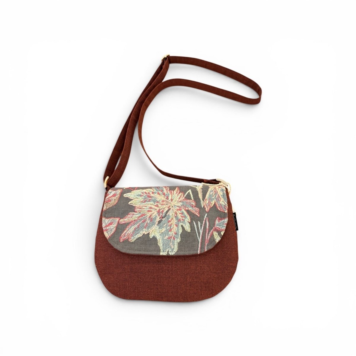 A small crossbody bag with a solid rust-brown body and strap, featuring a patterned floral front flap in muted green, pink, and beige tones, shown on a plain white background.