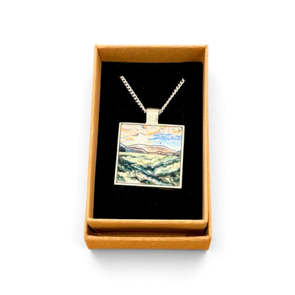 A square pendant necklace displayed in an open tan gift box with a black interior. The pendant features a colorful landscape painting with blue, green, yellow, and brown tones, suspended from a silver-toned chain.