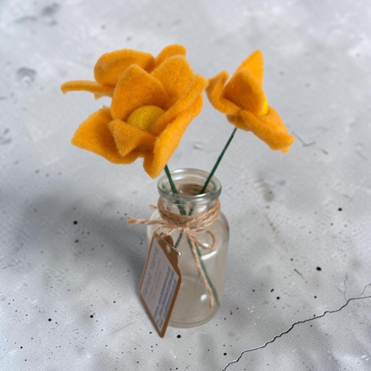 Three orange felt flowers in a small glass bottle on a textured white surface.
