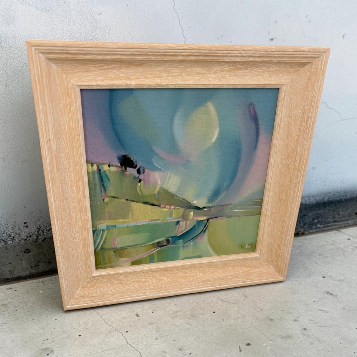 A square framed abstract painting leaning against a concrete wall on a concrete floor. The artwork features swirling brushstrokes of teal, green, yellow, and lavender, housed in a light natural oak frame.