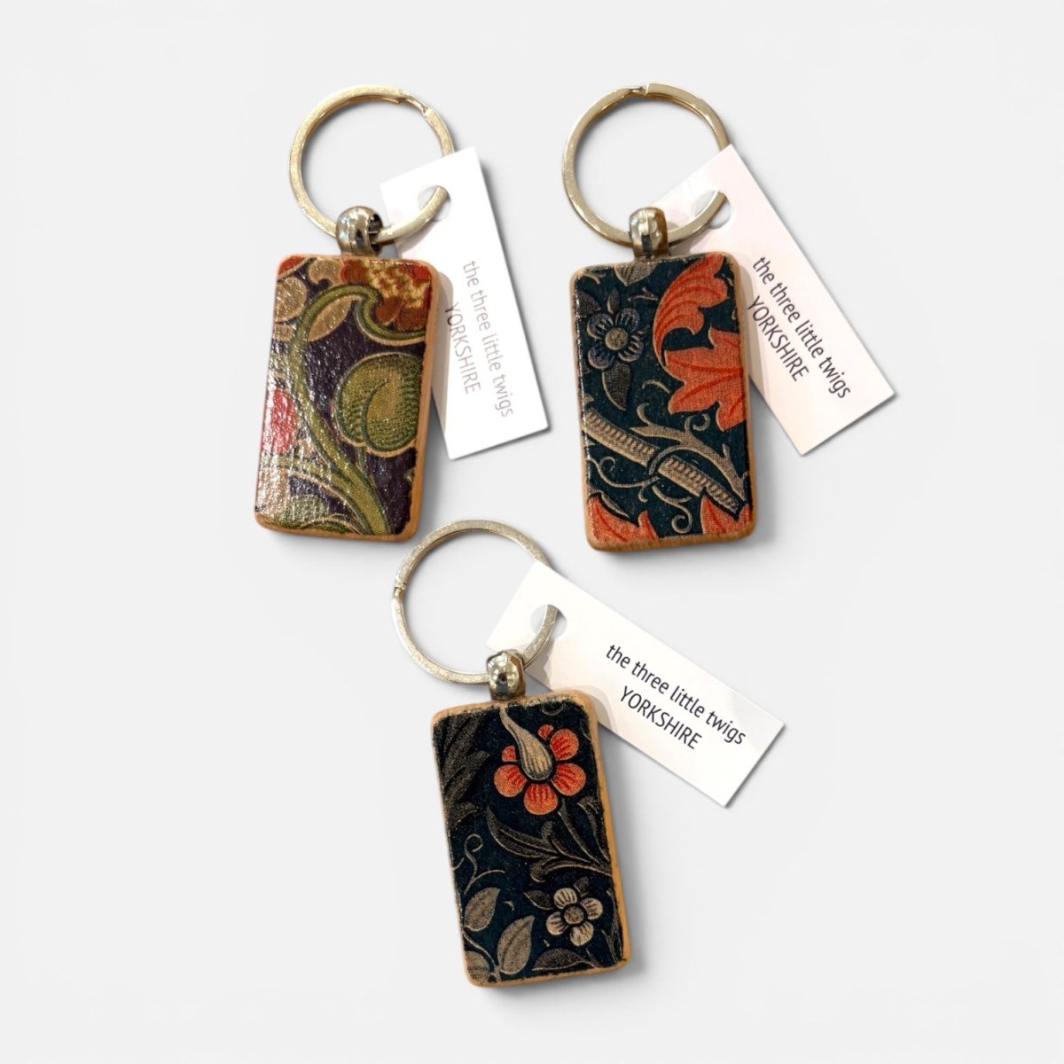 Three rectangular keychains with floral and ornamental patterns, each attached to a silver key ring and a white paper tag reading &