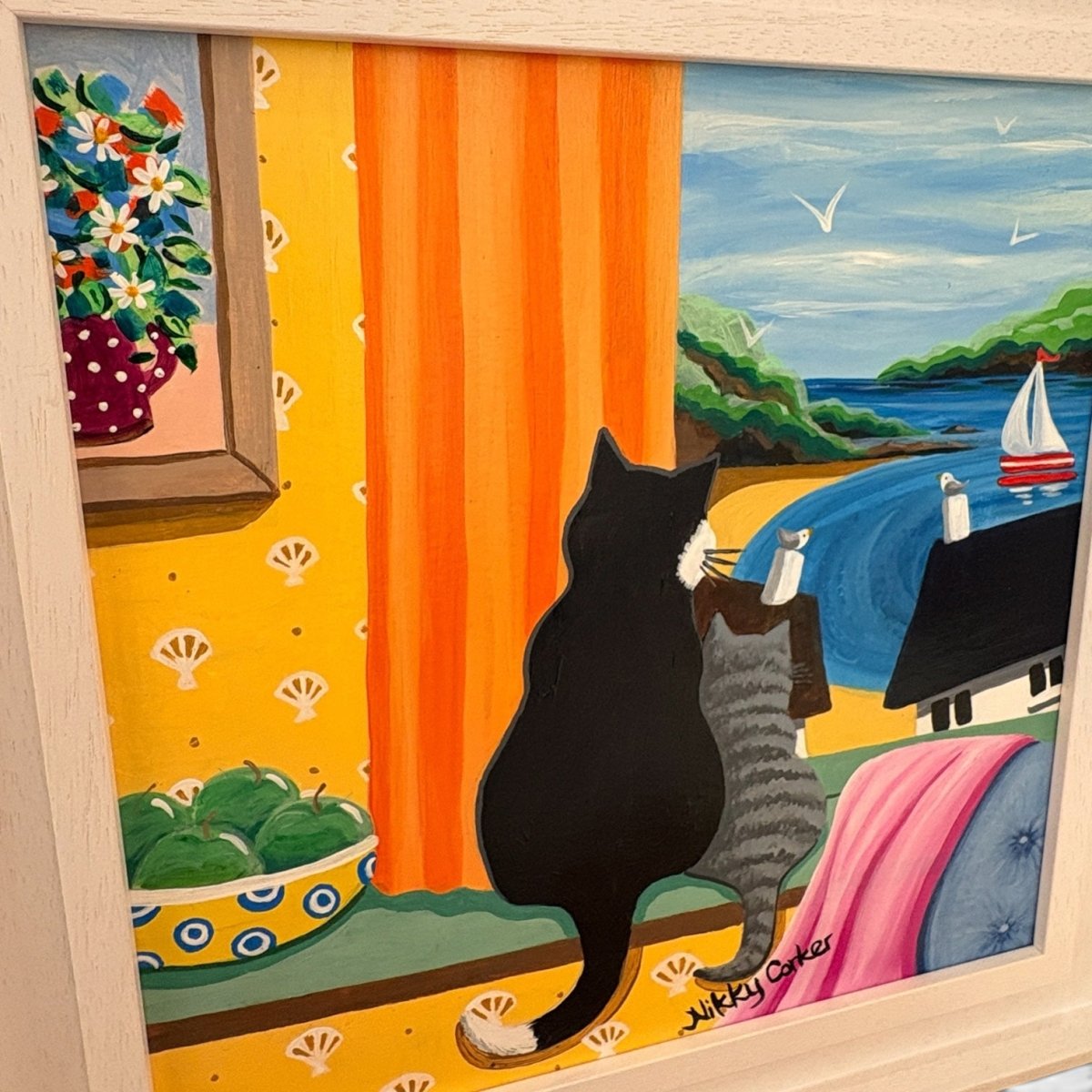 A framed colorful painting depicting a black cat sitting by a window looking out at a seaside beach scene, with an orange curtain, yellow patterned wall, bowl of green apples, and a sailboat on the water. Signed &