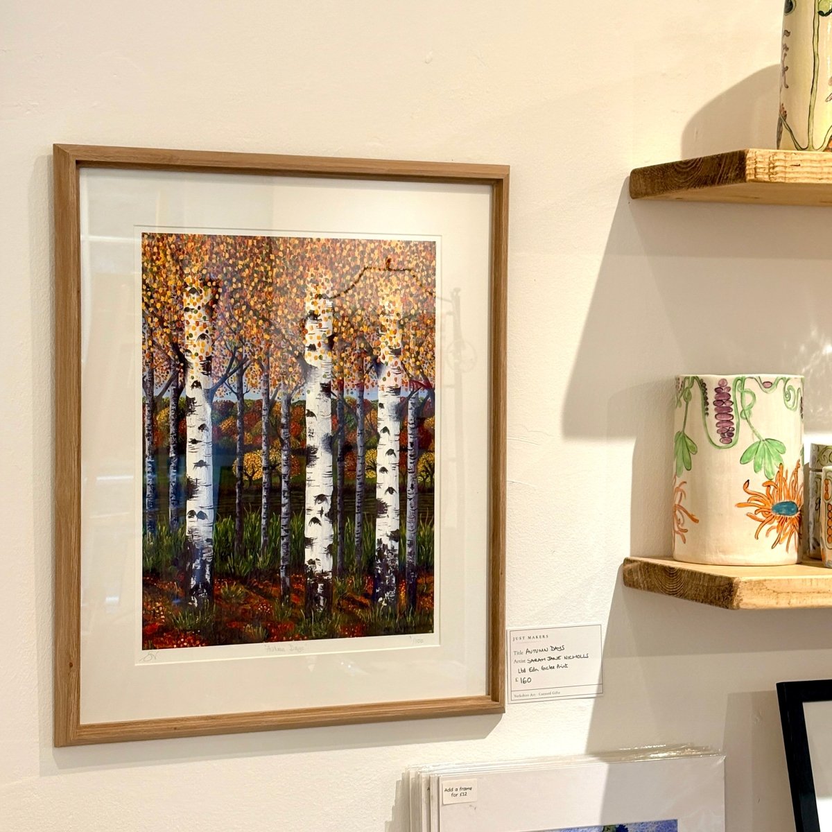 A framed art print depicting birch trees in an autumn forest, displayed on a white wall next to wooden shelves holding ceramic mugs.