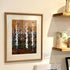A framed art print depicting birch trees in an autumn forest, displayed on a white wall next to wooden shelves holding ceramic mugs.