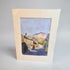 A square watercolor art print depicting a quaint village street with stone buildings, chimneys, and distant hills under a blue sky, mounted with a wide cream-colored mount on a neutral light gray background.