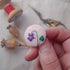 A hand holds a small round felt pin featuring an embroidered purple flower with a green stem and leaf. The background is softly blurred with sewing tools, including a spool of red thread, scissors, and wooden poppy pods.