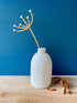 White vase with a dried plant against a blue wall on a wooden surface