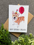 A greeting card featuring a watercolor-style corgi wearing a party hat and holding a red balloon, with the text &