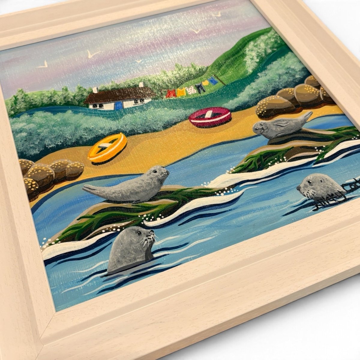 A framed hand-painted illustration of a coastal beach scene with seals in the water, a sandy shore, green hills, small boats, and a house. The artwork is shown at an angle inside a light natural wood frame on a white background.