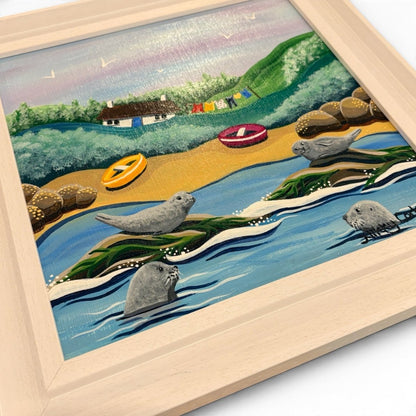 A framed hand-painted illustration of a coastal beach scene with seals in the water, a sandy shore, green hills, small boats, and a house. The artwork is shown at an angle inside a light natural wood frame on a white background.