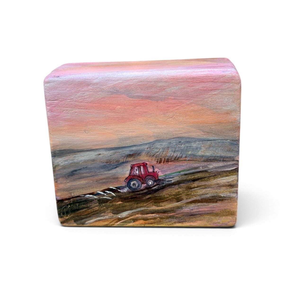A rectangular hand-painted wooden block featuring a landscape scene with a red tractor plowing a field under a pink and orange sunset sky, set against a plain white background.