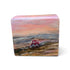 A rectangular hand-painted wooden block featuring a landscape scene with a red tractor plowing a field under a pink and orange sunset sky, set against a plain white background.