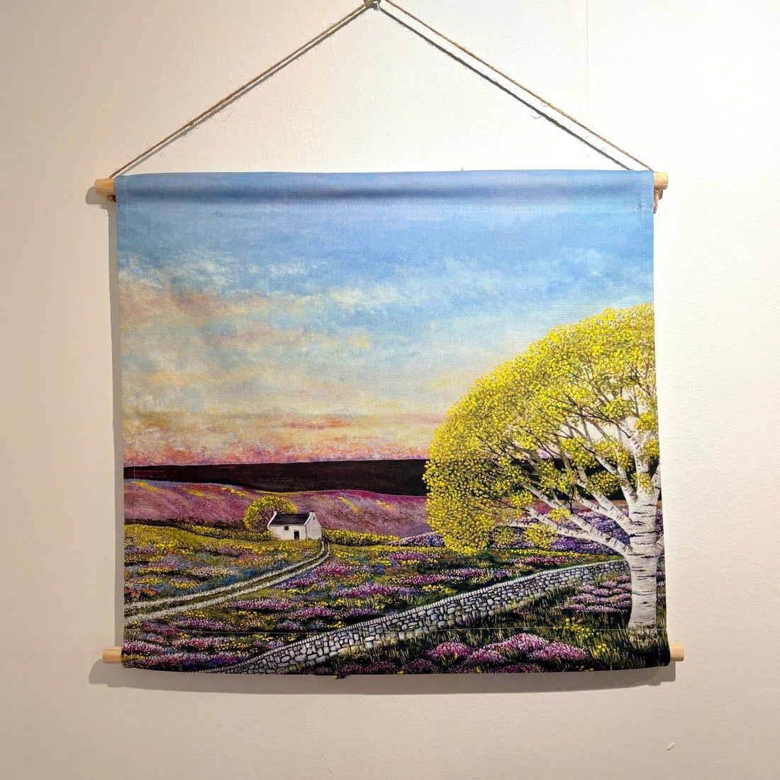A woven wall hanging tapestry depicting a rural landscape with a blue sky, purple flower fields, a small white cottage, and a large yellow-green tree, suspended from a wooden rod against a plain wall.